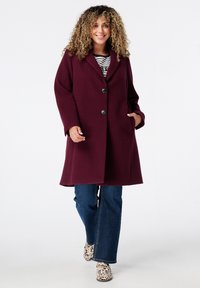 Manteau en laine bordeaux au design ajusté, à un bouton, avec poches latérales, porté sur un top rayé et un jean flare bleu.