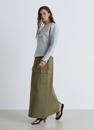Donna che cammina indossando una maglia a maniche lunghe grigio chiaro con catene decorative e una gonna cargo maxi verde oliva con sandali neri.