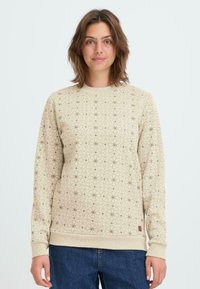 Sudadera beige con un patrón geométrico en marrón oscuro, puños y dobladillo acanalados, y un pequeño logo de tela en el borde inferior.