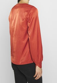 Blouse à manches longues de couleur rouille, fabriquée en tissu lisse. Elle présente une coupe décontractée, un col rond et des poignets froncés pour plus de détails.