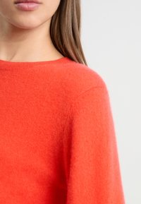 Pull rouge vif, texture douce, encolure ronde et design avec des épaules légèrement tombantes. Le matériau semble lisse avec peu de coutures visibles.