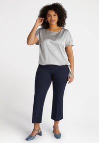 Top satin grigio chiaro con maniche corte, abbinato a pantaloni cropped blu. La modella indossa scarpe con tacco blocco blu e ha un'acconciatura riccia e texturizzata.