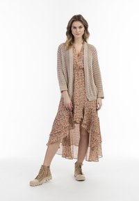 Beige gebreide cardigan over een bloemenjurk met een gerimpelde zoom. Lichtbruine enkellaarsjes met gestructureerde zolen. Glad, lichtgewicht materiaal.