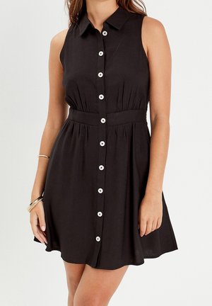 Femme portant une robe noire sans manches avec des boutons blancs à l'avant et une taille cintrée, accessoirisée avec des bracelets dorés sur un bras.