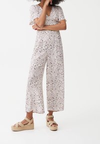 Bloemige jumpsuit met een V-hals, korte mouwen en wijde pijpen. Heeft een lichtroze achtergrond met paarse en zwarte accenten. Draagt met beige plateau-sandalen.