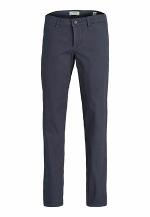 Enfant aux longs cheveux blonds, debout les bras croisés, portant une chemise beige à manches courtes avec des motifs, un t-shirt noir, un pantalon gris et des baskets beige.