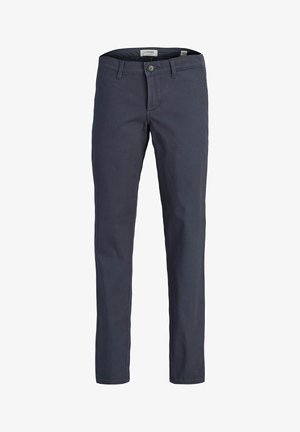Enfant aux longs cheveux blonds, debout les bras croisés, portant une chemise beige à manches courtes avec des motifs, un t-shirt noir, un pantalon gris et des baskets beige.