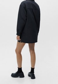 Personne portant une veste noire surdimensionnée, un short noir, des chaussettes noires et des chaussures à plateforme noires et chunky, se tenant contre un fond blanc uni.