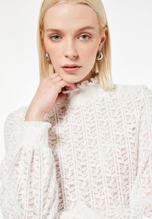 Kobieta o blond włosach, nosząca srebrne kolczyki koła i białą, teksturowaną koronkową bluzkę, opierająca rękę blisko brody.