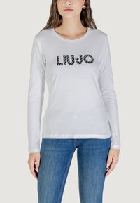 Modello che indossa una camicia bianca a maniche lunghe con la scritta "LIUJO" stampata sul davanti, abbinata a jeans blu. Sfondo neutro.