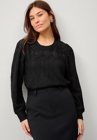 Blouse noire en dentelle à manches longues avec un motif texturé et un col festonné, associé à une jupe noire ajustée.