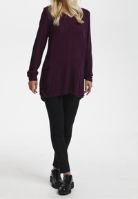 Långärmad, djup lila tunika med V-ringning och rundad nederkant, i kombination med svarta slim-fit byxor och kraftiga svarta skor.