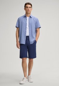 Camicetta azzurro chiaro a maniche corte con una finitura testurizzata, abbinata a pantaloni corti blu navy e sneakers bianche. Outfit semplice e informale.
