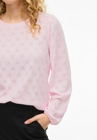 Woman wearing light pink long-sleeve blouse with eyelet pattern, paired with high-waisted black pants, hand in pocket.