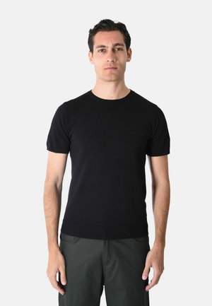 Giovane uomo con capelli corti e scuri che indossa una maglietta nera aderente a maniche corte e pantaloni scuri, in piedi davanti a uno sfondo chiaro e semplice.