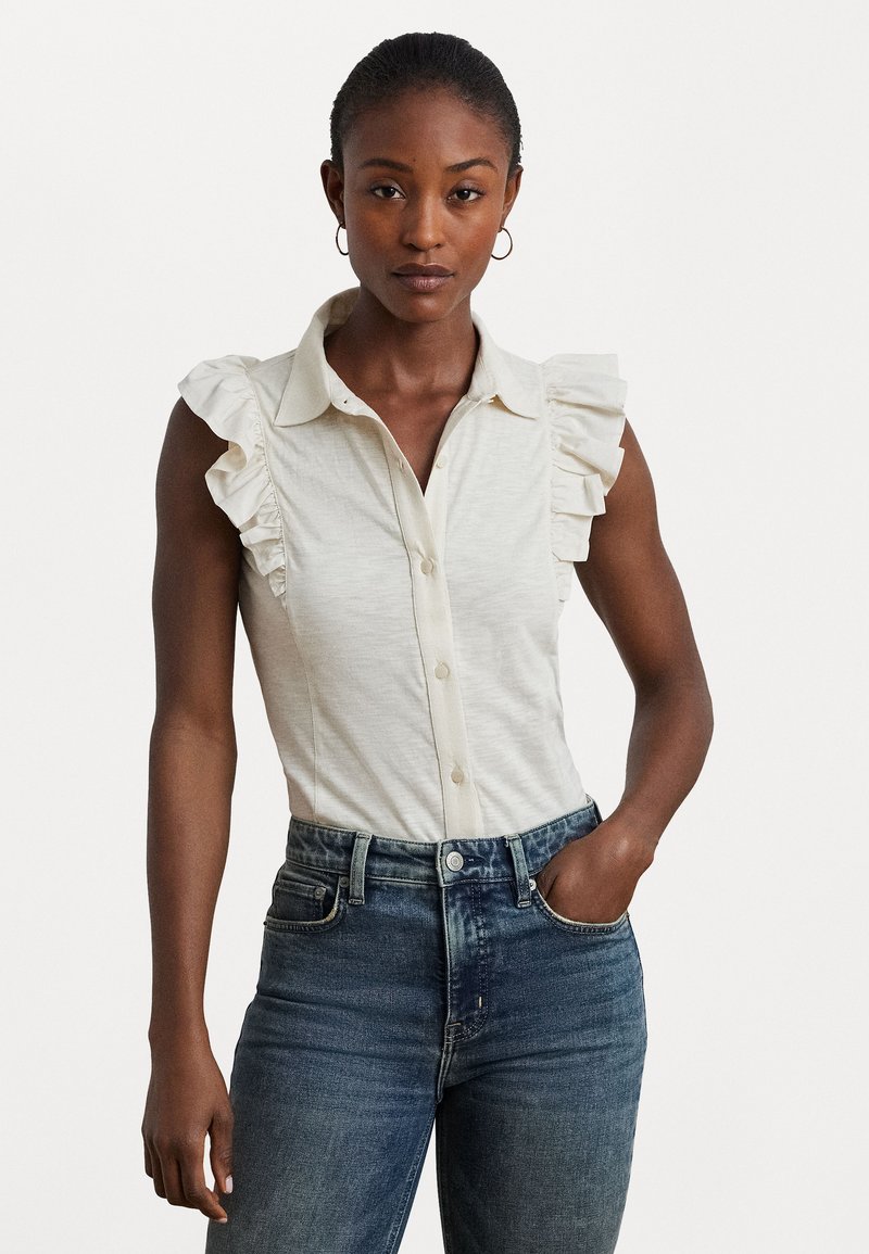 Blouse sans manches de couleur beige clair avec des accents de volants le long des épaules, fermeture à boutons et une texture douce, associée à un jean en denim bleu.