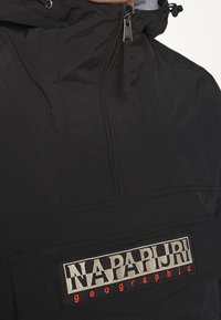 Chaqueta negra de pullover con cuello alto, cremallera y un bolsillo frontal tipo pouch; presenta un logo rectangular con texto en blanco y naranja.