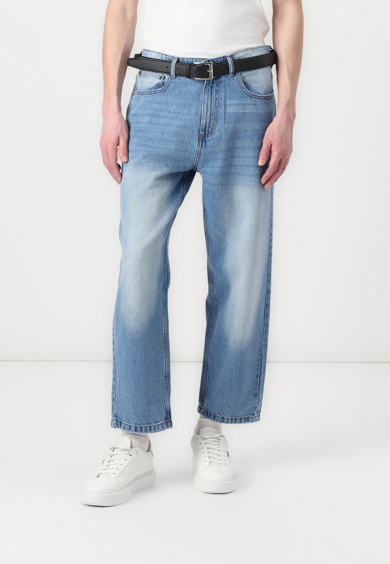 Jeans de mezclilla azul claro con corte recto, con secciones descoloridas, un cinturón negro y zapatillas blancas. Diseño simple y corte clásico.