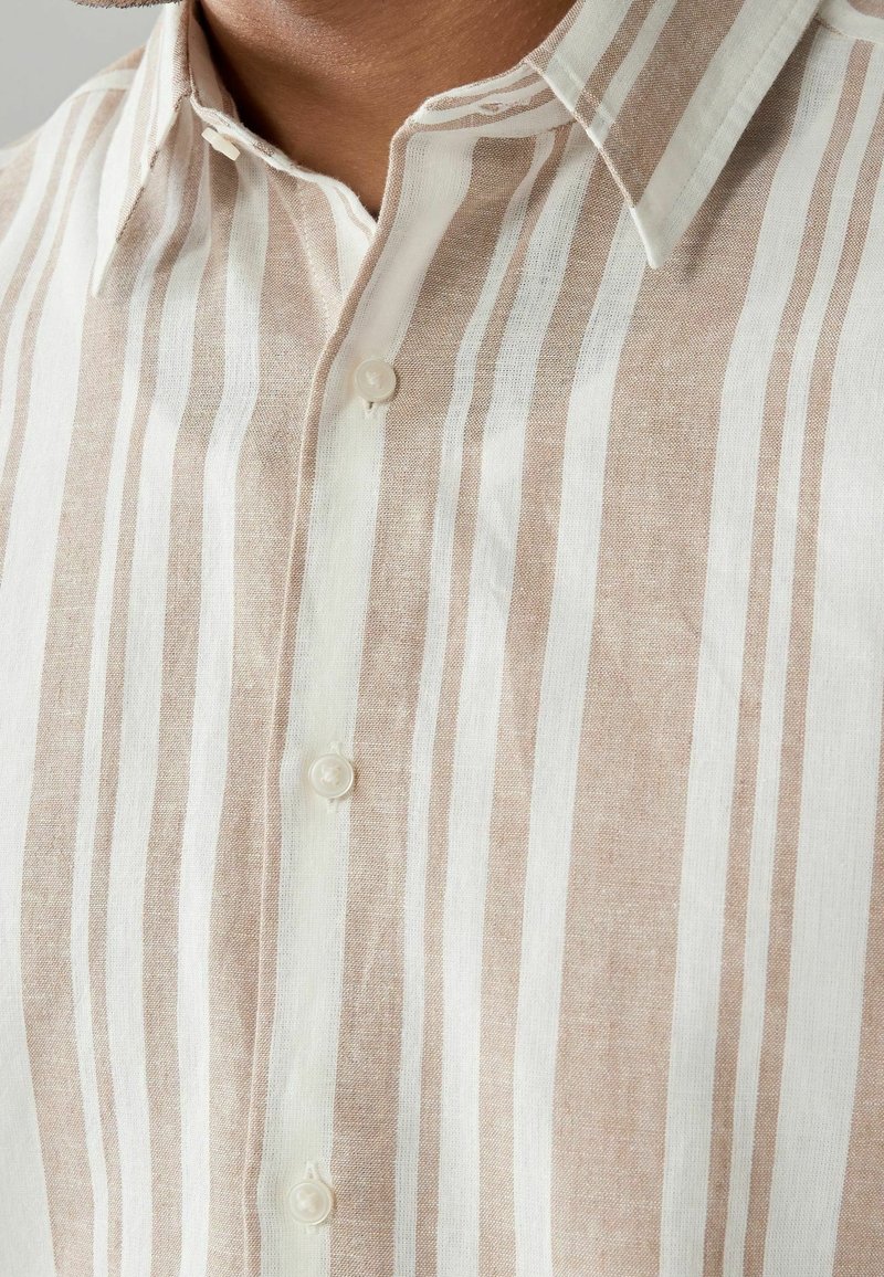 Chemise à col avec des rayures verticales beige et blanches, partiellement boutonnée, montrant un tissu texturé près du cou et de la poitrine.