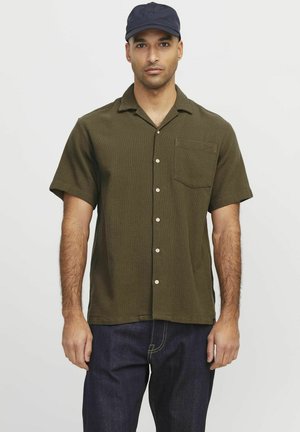 Chemise vert olive à manches courtes, à boutons, en tissu texturé, avec une poche poitrine unique et des boutons crème, associée à un jean foncé.