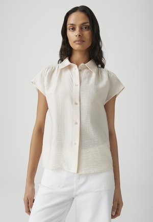 Frau mit langen dunklen Haaren, die ein cremefarbenes Kurzarmhemd mit Knöpfen und weiße Hosen vor einem einfarbigen hellen Hintergrund trägt.