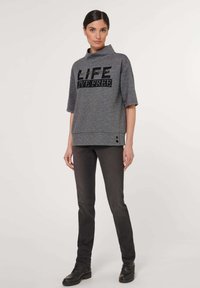 Grijze geblokte blouse met hoge kraag, voorzien van een zwarte "LIFE LIVE FREE" grafische print, gecombineerd met donkergrijze slim-fit broek en zwarte laarzen.