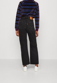 Jeans larghi neri in denim, con tasche posteriori, dettagli di cucitura e una cintura etichettata. Indossati con un maglione a righe e sneaker bianche.
