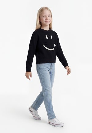 Blondína chodí, má na sebe čierny sveter s bielym smajlíkom, svetlomodré rifle a biele tenisky.