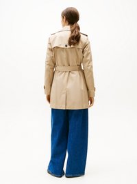 Donna con capelli castani raccolti in una coda di cavallo, che indossa un trench beige con cintura e jeans ampi blu, in piedi davanti a uno sfondo bianco.