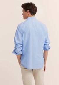 Mann steht mit dem Rücken zur Kamera, trägt ein hellblaues Langarmshirt mit hochgekrempelten Ärmeln und beigen Hosen.