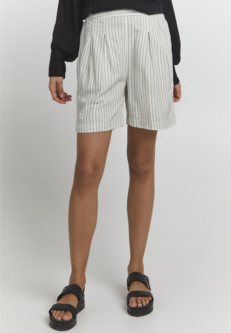 Shorts légers de couleur crème avec des rayures verticales noires, présentant un devant plissé et une coupe décontractée. Portés avec des sandales plates noires.