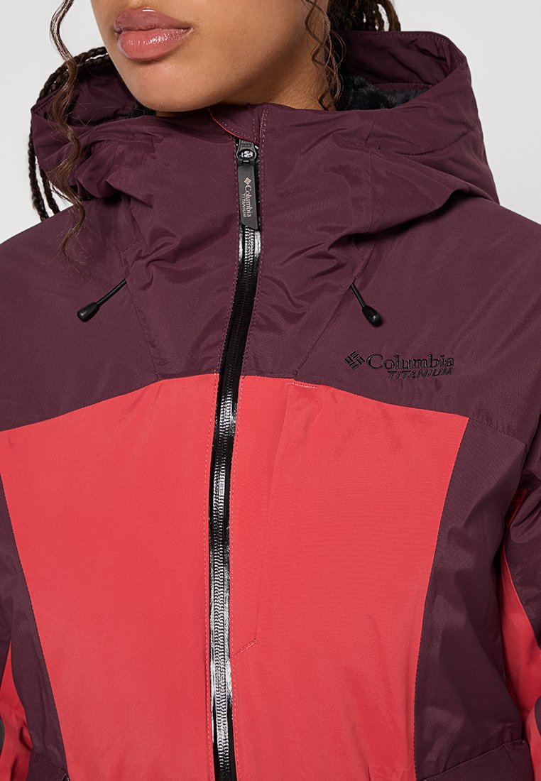 Kastanienbraune und leuchtend rote Jacke mit einem Reißverschluss vorne, verstellbare Kapuze und schwarzen Akzenten. Columbia-Logo auf der Vorderseite.