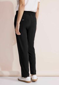 Zwarte broek met een elastische tailleband, recht gesneden en met achterzakken. Draag met witte schoenen tegen een neutrale achtergrond.