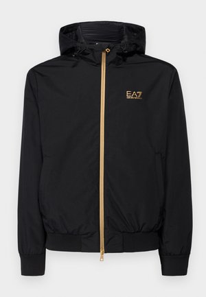 Black hooded jacket made of lightweight material with a gold zipper and logo accent. Features elastic cuffs and a fitted waist.