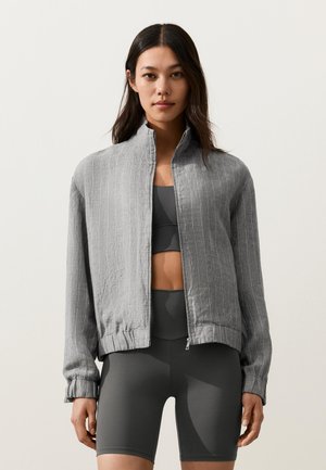 Frau trägt eine hellgraue Zip-Up-Jacke über einem dunkelgrauen Sport-BH und passenden hoch taillierten Fahrradhosen, steht vor einem schlichten Hintergrund.