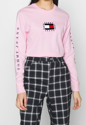 Osoba nosząca jasnoróżową koszulkę Tommy Jeans z długim rękawem z logo na piersi i rękawach, w połączeniu z czarnymi i białymi spodniami w kratę.