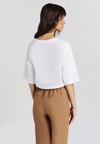 Donna con capelli scuri che indossa un top bianco a maniche corte e morbido, e pantaloni marroni a vita alta, rivolta di lato su uno sfondo semplice.