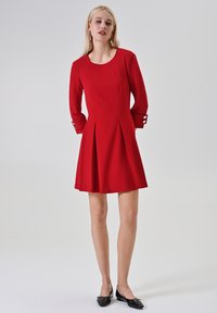 Rode jurk met lange mouwen, voorzien van een ronde halslijn, plooien en knoopaccenten op de mouwen. Gecombineerd met zwarte platte schoenen.