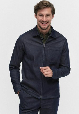 Man smiling and wearing a dark blue zip-up jacket over an olive-green shirt with matching dark blue pants against a plain background.