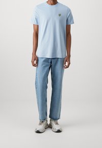 Jasnoniebieska bawełniana koszulka z okrągłym dekoltem i małym haftowanym logo na piersi, zestawiona z luźnymi jasnymi dżinsami.