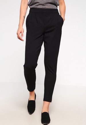 Osoba ubrana w czarne, dopasowane spodnie z bocznymi kieszeniami i czarne wsuwane buty, stojąca na jednolitym jasnym tle.