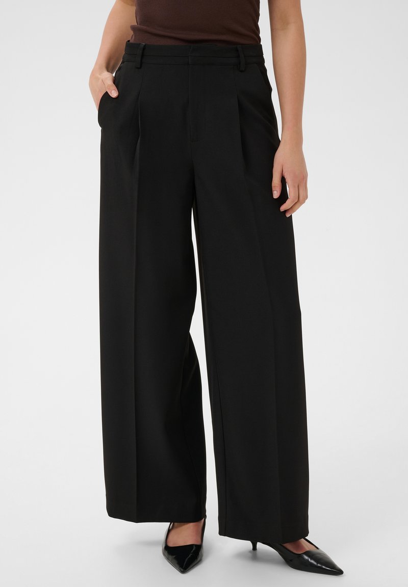 Femme portant un pantalon noir taille haute à jambes larges avec des plis, associé à des talons noirs pointus et un haut sans manches marron foncé.