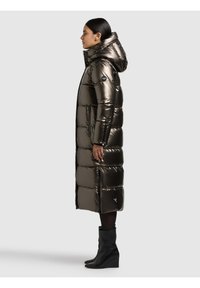 Duga, tamna metalik jakna s punjenjem, s kapuljačom, prošivenim jastučićima, bočnim patentnim zatvaračima i oznakom na lijevoj ruci. Nošena s crnim čizmama.