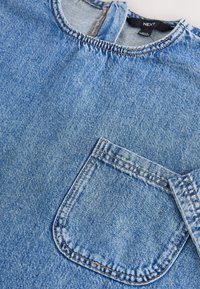 Camicia di denim azzurro chiaro con collo rotondo, maniche corte e tasca frontale. Presenta dettagli cuciti e una texture sbiadita.
