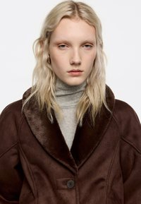 Osoba o blond włosach, ubrana w brązowy płaszcz z futrzanym kołnierzem na jasnoszarym golfie, zwrócona przodem, z neutralnym wyrazem twarzy, na białym tle.