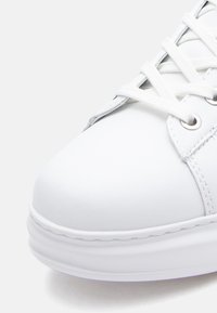 White sneaker made of smooth synthetic material with a rounded toe, flat sole, and woven white laces. Features silver eyelets and stitching.