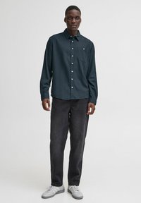 Camicia verde scuro con bottoni, maniche lunghe e taschino sul petto, abbinata a jeans neri e sneakers bianche. Tessuto semplice, vestibilità casual.