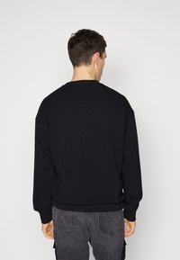 Sudadera negra de tejido suave, con cuello redondo y puños acanalados. Combinada con pantalones cargo grises, vista desde atrás.
