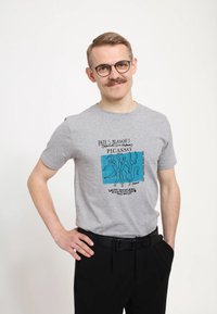 Šedé tričko s krátkým rukávem s abstraktním modrým designem a černým textem, spárované s černými kalhotami a černým páskem.