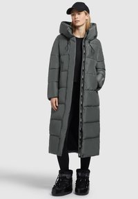 Lang grå puffjakke med hætte, med front lynlås, elastiske snore og sidelommer. Sammen med sorte støvler og en kasket.